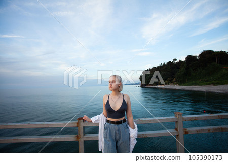 Cute blonde woman in white shirt and tank top standing on pier on the background of sea 105390173