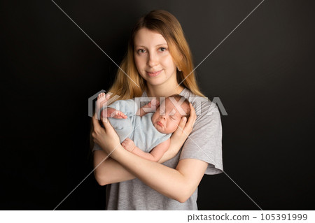 A cute newborn baby boy sleeps in a blue overalls in the first days of life. In the arms of the mother. Warm motherly hugs. On a black background. Professional portrait photography. 105391999