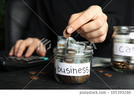 Woman putting coin into jar with money and tag BUSINESS on black table, closeup 105393280