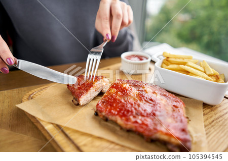 Lunch in a restaurant, a woman cuts Delicious Pork ribs. Full rack of ribs BBQ on wooden plate with french fries and salad Lunch in a restaurant, a woman cuts Delicious Pork ribs. Full rack of ribs BBQ on wooden plate with french fries and salad 105394545