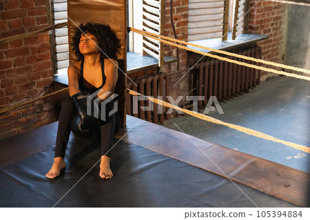 Woman fighter girl power. African american woman fighter with boxing gloves sitting on boxing ring waiting and resting after fight. Strong powerful girl in gym. Strength fit body workout training Woman fighter girl power. African american woman fighter with boxing gloves sitting on boxing ring waiting and resting after fight. Strong powerful girl in gym. Strength fit body workout training 105394884