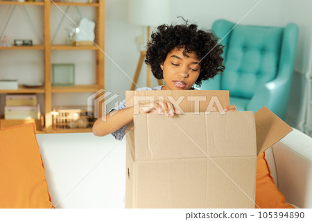 African girl unpacking delivery looking in box. Happy woman opening carton box. Female getting parcel looking at delivered goods items. Satisfied client positive feedback quick express deliver African girl unpacking delivery looking in box. Happy woman opening carton box. Female getting parcel looking at delivered goods items. Satisfied client positive feedback quick express deliver 105394890