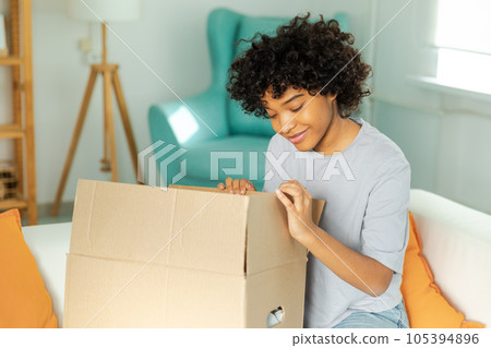 African girl unpacking delivery looking in box. Happy woman opening carton box. Female getting parcel looking at delivered goods items. Satisfied client positive feedback quick express deliver African girl unpacking delivery looking in box. Happy woman opening carton box. Female getting parcel looking at delivered goods items. Satisfied client positive feedback quick express deliver 105394896