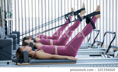 Three Asian women in pink sportswear doing pilates exercises with a reformer bed.  105398800