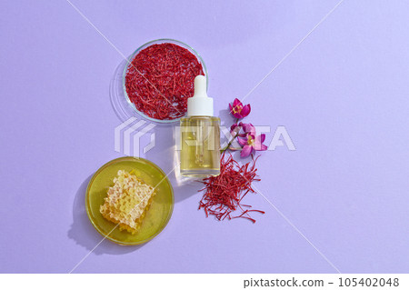 Fresh ingredients saffron and honey displayed on purple background with unbranded serum bottle. Scene for advertising. With many uses, saffron has become a great medicine, both for health and beauty. 105402048