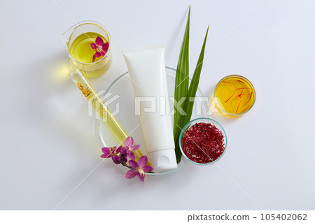 Top view of a white plastic tube mockup for cosmetic displayed on a petri dish with stigmas saffron, fresh crocus sativus, green leaves decorated with glassware filled liquid on a white background 105402062