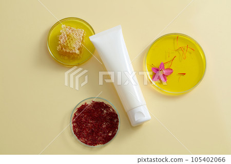 Against the beige background, a white cosmetic tube decorated with petri dishes containing stigmas saffron, honey and yellow liquid. Advertising scene for cosmetic with natural extract Against the beige background, a white cosmetic tube decorated with petri dishes containing stigmas saffron, honey and yellow liquid. Advertising scene for cosmetic with natural extract 105402066