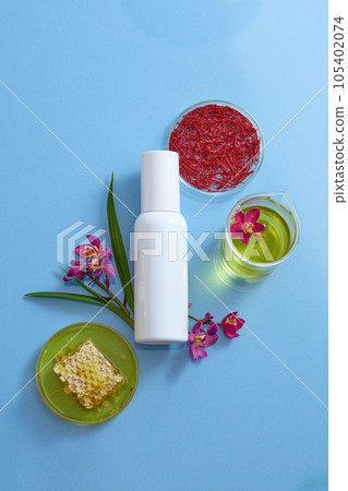 Advertising photo for cosmetic with ingredient from stigmas saffron and honey. A white bottle unbranded decorated with stigmas saffron, fresh crocus sativus and beeswax on blue background 105402074