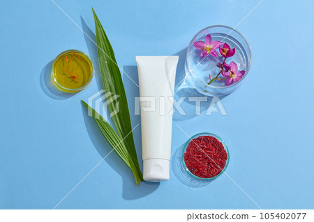 Top view of a white cosmetic bottle container cream or facial cleanser decorated with fresh crocus sativus, stigmas saffron and green leaves on blue background. 105402077