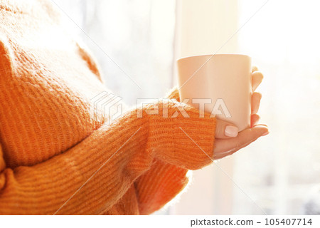 Woman in orange sweater holding cup of tea or coffee near window at home Woman in orange sweater holding cup of tea or coffee near window at home 105407714