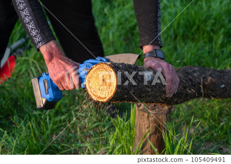 Hand-held portable camping electric saw on a battery for cutting firewood and wood. Close-up, a hand sawing a log, splinters flying Hand-held portable camping electric saw on a battery for cutting firewood and wood. Close-up, a hand sawing a log, splinters flying 105409491