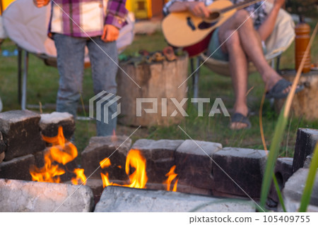 A girl in a plaid shirt roasts marshmallows on a fire in the yard of the house, father plays the guitar. Evening family get-together by the campfire, outdoor picnic in tourist chairs A girl in a plaid shirt roasts marshmallows on a fire in the yard of the house, father plays the guitar. Evening family get-together by the campfire, outdoor picnic in tourist chairs 105409755