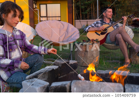 A girl in a plaid shirt roasts marshmallows on a fire in the yard of the house, father plays the guitar. Evening family get-together by the campfire, outdoor picnic in tourist chairs A girl in a plaid shirt roasts marshmallows on a fire in the yard of the house, father plays the guitar. Evening family get-together by the campfire, outdoor picnic in tourist chairs 105409759