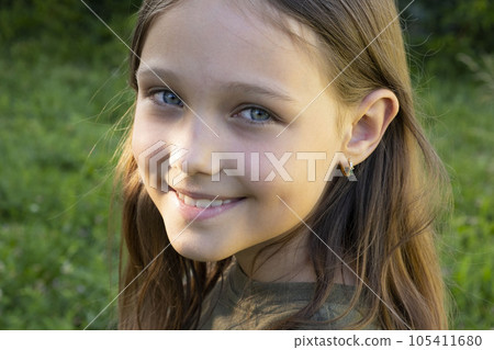 Portrait of a beautiful brunette girl looking at the camera and smiling, standing on the street against the backdrop of greenery on a summer day. Young curious child raises his eyes and lowered his 105411680