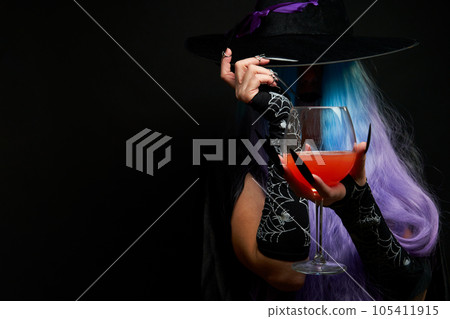 Witch with blue and violet hair, black hat with violet ribbon and long black nails is holding wine glass in her hands. 105411915