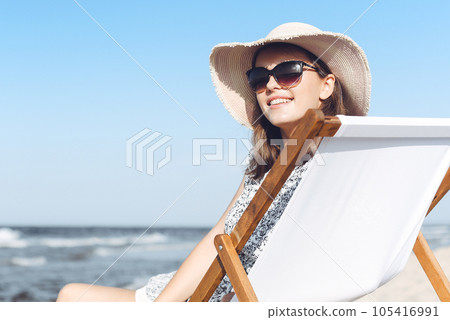 Happy brunette woman wearing sunglasses and hat relaxing on a wooden deck chair at the ocean beach Happy brunette woman wearing sunglasses and hat relaxing on a wooden deck chair at the ocean beach 105416991