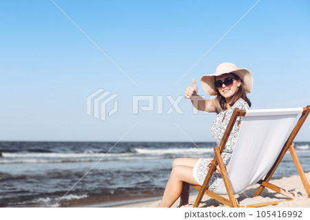 Happy brunette woman sitting on a wooden deck chair at the ocean beach while showing Thumb up Happy brunette woman sitting on a wooden deck chair at the ocean beach while showing Thumb up 105416992