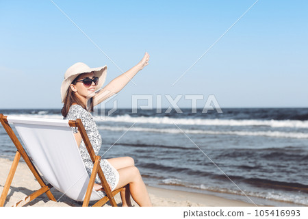 Happy brunette woman sitting on a wooden deck chair at the ocean beach while waving and greeting somebody with her hand Happy brunette woman sitting on a wooden deck chair at the ocean beach while waving and greeting somebody with her hand 105416993