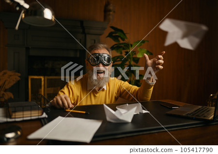 Funny overjoyed mature man wearing pilot goggles playing with origami plane Funny overjoyed mature man wearing pilot goggles playing with origami plane 105417790