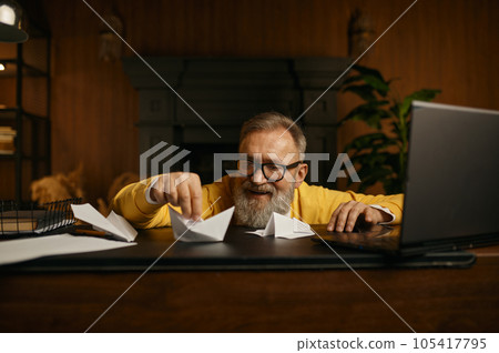 Happy excited mature grey-haired man playing with paper boat at workplace Happy excited mature grey-haired man playing with paper boat at workplace 105417795