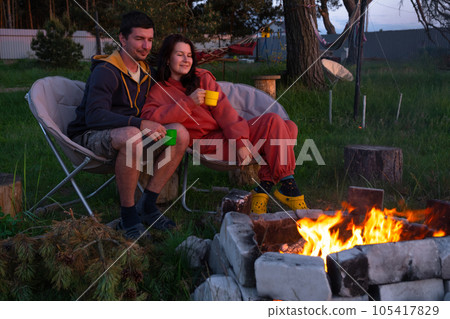 Couple in love is sitting by the outdoor campfire in the courtyard of the house on camping chairs, a romantic evening. a man and a woman warm their hands by the fire, drink tea 105417829