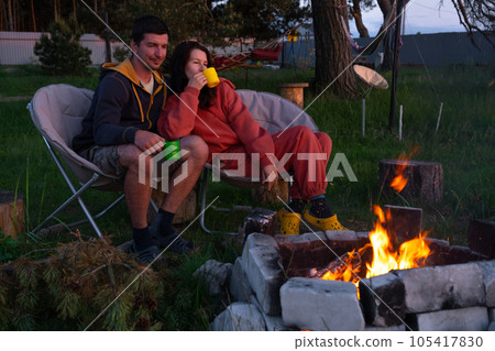 Couple in love is sitting by the outdoor campfire in the courtyard of the house on camping chairs, a romantic evening. a man and a woman warm their hands by the fire, drink tea Couple in love is sitting by the outdoor campfire in the courtyard of the house on camping chairs, a romantic evening. a man and a woman warm their hands by the fire, drink tea 105417830