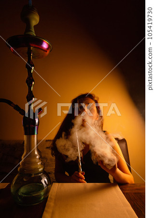 Relaxing on the couch, the woman smokes a hookah and releases a cloud of steam. Orange tint Relaxing on the couch, the woman smokes a hookah and releases a cloud of steam. Orange tint 105419730