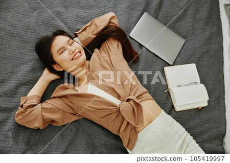 Young female in casual wear smiling with closed eyes while lying on comfortable bed with opened laptop and notebook near her. 105419997