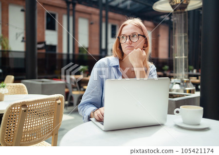 Stressed young blonde woman using laptop at cafe, sitting at table with notepad and cup of coffee alone Stressed young blonde woman using laptop at cafe, sitting at table with notepad and cup of coffee alone 105421174
