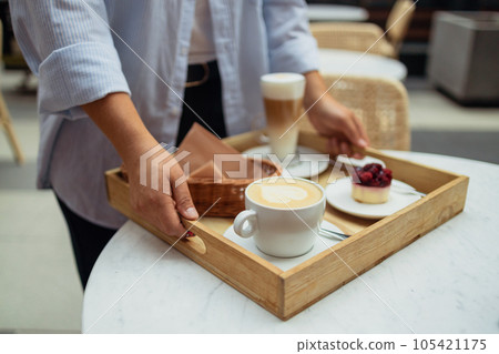 Happy smiling handsome woman waiter in a restaurant holding a tray with a latte coffee, cappuccino and berry cake in a restaurant Happy smiling handsome woman waiter in a restaurant holding a tray with a latte coffee, cappuccino and berry cake in a restaurant 105421175