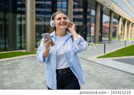 Smiling caucasian young business woman listening to the podcast e-book music song singer rock band in headphones earphones, choosing sound track on cellphone walking on urban city street. High quality 105421210