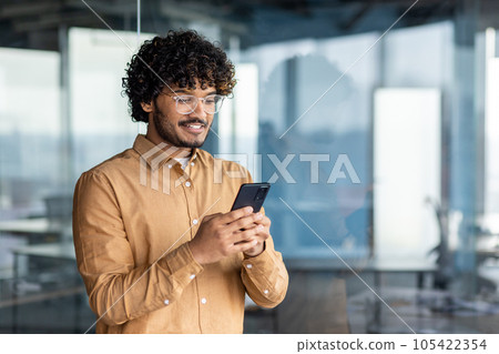 Hispanic man smiles and looks at the phone screen, businessman inside the office holds a smartphone, browses online internet pages, makes a call, and uses an application, near the window in a shirt. 105422354