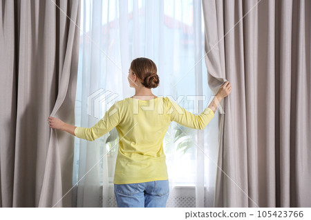 Woman opening window curtains at home in morning Woman opening window curtains at home in morning 105423766