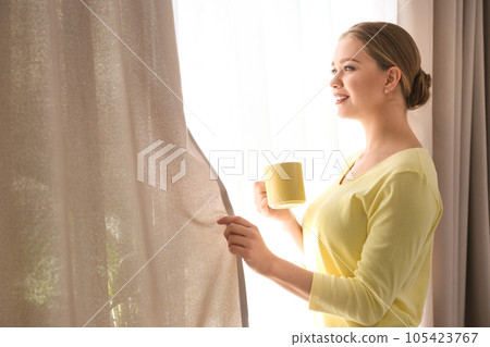 Woman with cup of coffee opening window curtain at home in morning Woman with cup of coffee opening window curtain at home in morning 105423767