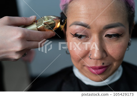 The hairdresser shaves the temple of a female client. Asian woman with short pink hair in barbershop. 105429173