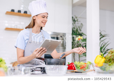 Beautiful model happy woman healthy food cooking vegetable salad in kitchen with online tablet Beautiful model happy woman healthy food cooking vegetable salad in kitchen with online tablet 105429543