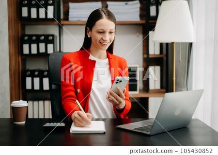 Confident business expert attractive smiling young woman typing laptop ang holding digital tablet on desk in office.. Confident business expert attractive smiling young woman typing laptop ang holding digital tablet on desk in office.. 105430852