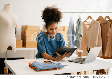 Calm curly brunette dark skinned woman on desk in modern office of fashion designer and holds tablet and smartphone. Calm curly brunette dark skinned woman on desk in modern office of fashion designer and holds tablet and smartphone. 105434705
