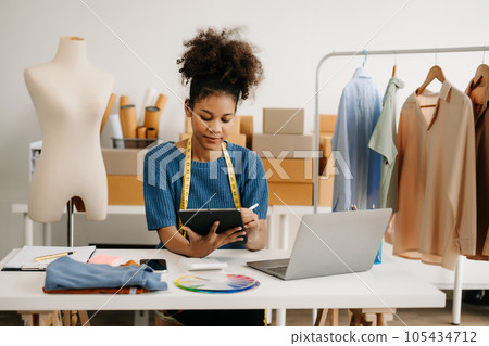 Calm curly brunette dark skinned woman on desk in modern office of fashion designer and holds tablet and smartphone. 105434712