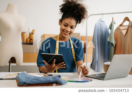 Calm curly brunette dark skinned woman on desk in modern office of fashion designer and holds tablet and smartphone. 105434713