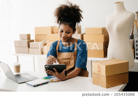 Startup small business SME, Entrepreneur owner African woman using smartphone or tablet taking receive and checking online purchase shopping order to preparing pack product box. in office. Startup small business SME, Entrepreneur owner African woman using smartphone or tablet taking receive and checking online purchase shopping order to preparing pack product box. in office. 105435066