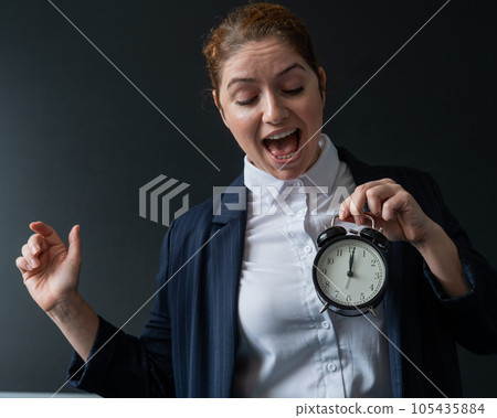 Happy female teacher in a pantsuit looks at the alarm clock while standing at the blackboard. A woman rejoices at the end of the lessons at school. Happy female teacher in a pantsuit looks at the alarm clock while standing at the blackboard. A woman rejoices at the end of the lessons at school. 105435884