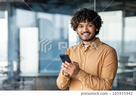 Portrait of a young businessman inside the office, hispanic man is smiling and looking at the camera, the man is holding a phone in his hands, typing a message and browsing the internet. Portrait of a young businessman inside the office, hispanic man is smiling and looking at the camera, the man is holding a phone in his hands, typing a message and browsing the internet. 105435942