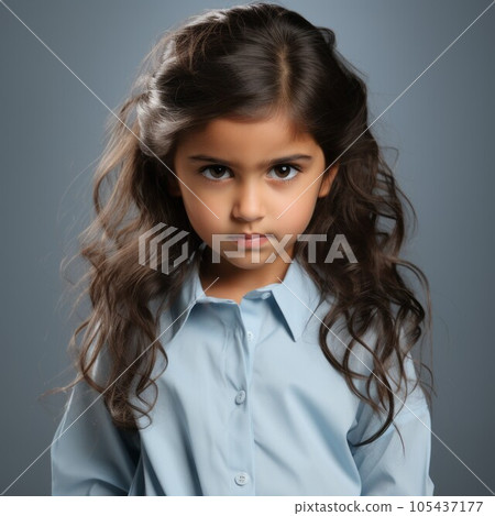 Portrait of an angry little girl with wavy brown hair. Closeup face of a furious Caucasian child on a blue background. White European small kid with no expression in a blue shirt looking at camera. 105437177