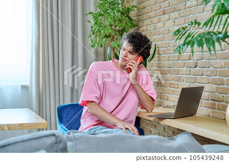 Young male talking on phone, sitting at desk at home with laptop 105439248