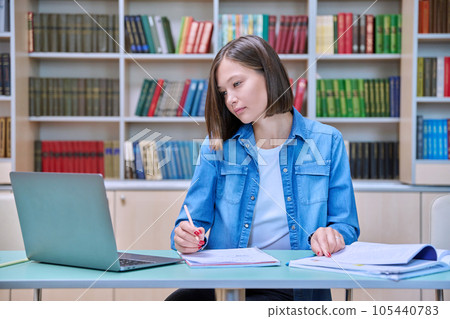 Young female university student studying inside library, using laptop, writing in notebook 105440783