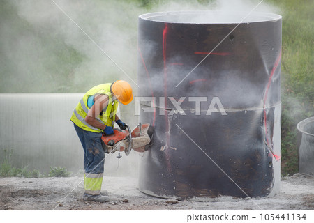 Reconstruction of the road in the city. A worker cuts holes in a concrete barrel to collect rainwater installed under the pavement. Use of a helmet and respirator, compliance with safety regulations. Reconstruction of the road in the city. A worker cuts holes in a concrete barrel to collect rainwater installed under the pavement. Use of a helmet and respirator, compliance with safety regulations. 105441134