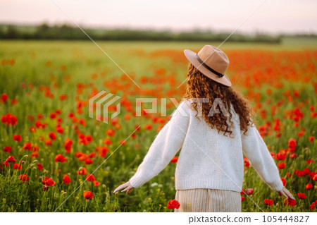Back view. Young curly woman in hat posing in the poppy field. Summer landscape. 105444827