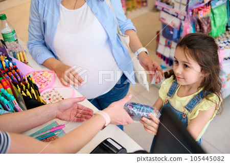 Top view little girl holding out a pencil case to a saleswoman, standing at the counter while buying school stationery 105445082