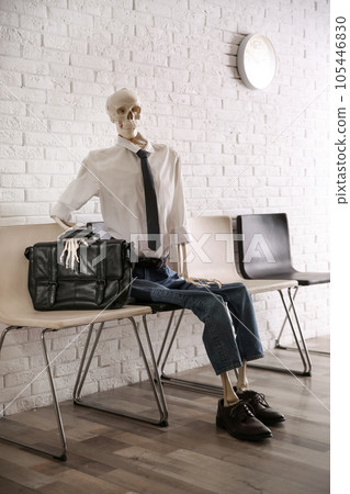 Human skeleton in office wear sitting on chair near brick wall indoors 105446830